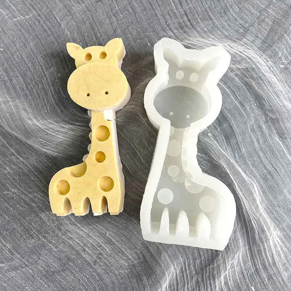 A giraffe-shaped silicone mould displayed on a textured surface. The left mould is made of a pale yellow material with circular holes and defined features, while the right mould is white and clear, showing the same giraffe shape. The design includes a long neck, rounded head, and spots along the body.