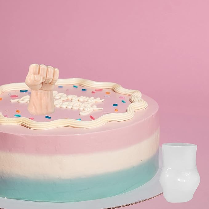 A colorful three-layered cake with pink, white, and teal frosting sits against a pink background. Atop the cake is a small, pink decorative fist made from silicone, surrounded by white decorations and colorful sprinkles. Next to the cake is a small, white, opaque cup.