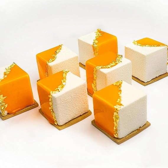 A collection of six square desserts arranged on a white surface. Each dessert features a unique design with a bright orange glossy top and a textured white base, partially covered by a thin strip of gold detailing along one edge. The desserts are presented on small square golden bases.