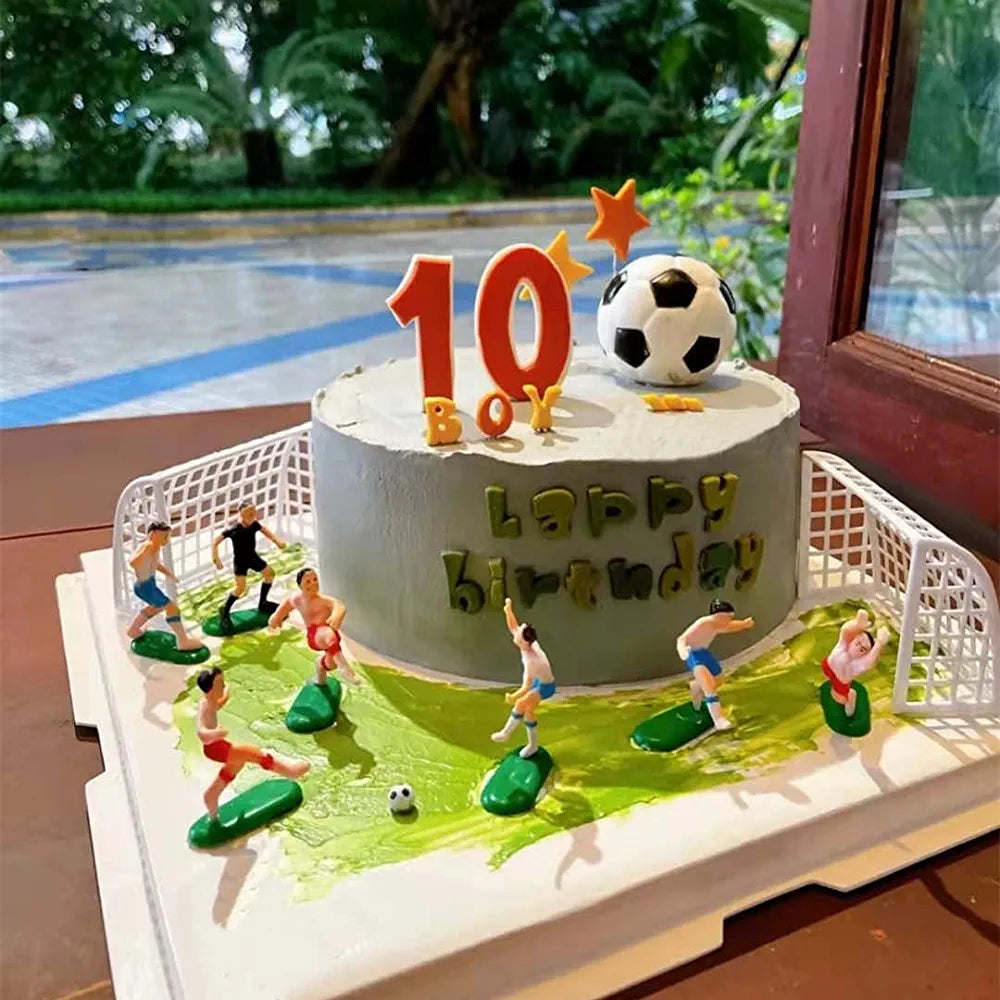 A birthday cake designed with a soccer theme. The cake features a circular gray frosting layer adorned with the words &quot;Happy birthday&quot; in colorful lettering. Atop the cake is a soccer ball and the number &quot;10&quot; in orange with a star. Miniature soccer player figurines in different poses are placed around the cake, depicting a soccer match, with a soccer goal and green turf-like frosting. The scene is set against a backdrop of greenery.