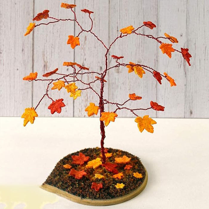 Image description for accessibility: A decorative miniature tree with branches made of thin wire, adorned with small maple leaves in various autumn colors, including red, orange, and yellow. The tree is positioned on a circular base covered with brown material resembling soil, also scattered with additional small leaves in similar colors. The background features a light wooden panel.