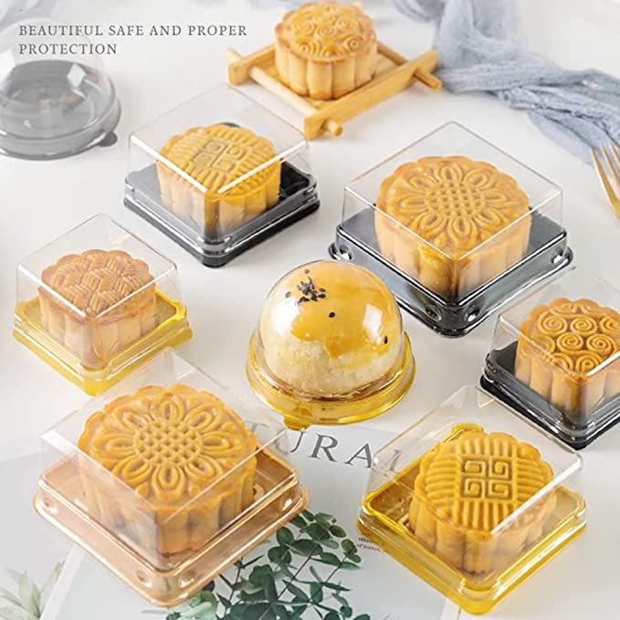 This image features a collection of mini mooncakes displayed in transparent boxes. There are several mooncakes with intricate designs, each contained in individual plastic packaging, including varying colors like clear, black, and yellow. One mooncake is slightly larger and round, topped with a small decoration, while another is positioned on a small wooden tray. The background includes soft, neutral tones with some greenery peeking through, enhancing the presentation.