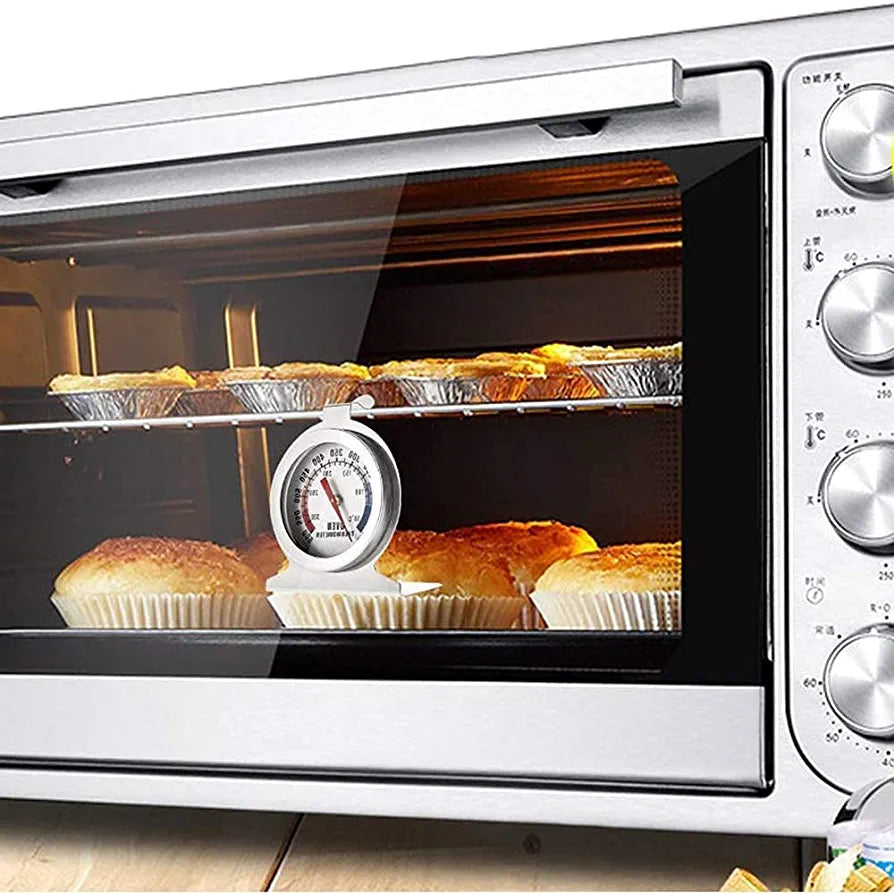 An oven with a glass door is shown, displaying several muffins baking on two shelves inside. In the foreground, there is an analog oven thermometer sitting on the shelf, showing temperature readings. The oven has a stainless steel finish and control knobs on the right side. The muffins appear golden brown, and the overall scene conveys a warm, inviting atmosphere for baking.