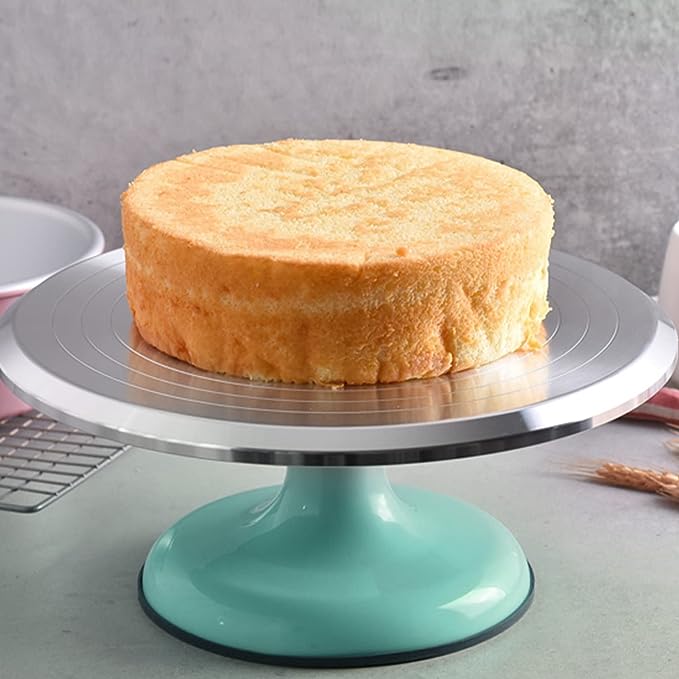 A round, golden sponge cake is placed on an aluminum revolving turntable. The turntable features a shiny silver surface and a light teal base. In the background, there are other baking tools, including a pink bowl and a wire cooling rack, along with some decorative items like wheat stalks. The setting has a modern kitchen aesthetic with a neutral gray backdrop.