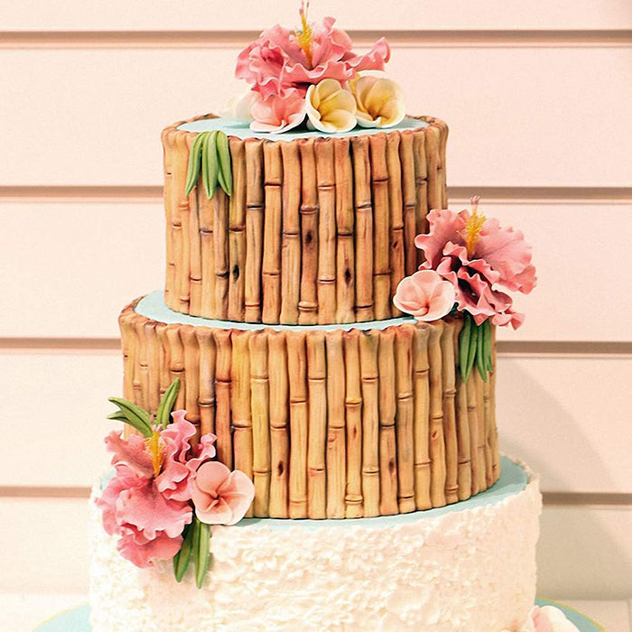 A two-tiered cake decorated to resemble bamboo, featuring realistic bamboo detailing along the sides. The cake is adorned with various colorful flowers, including pink and yellow blooms, and green leaf accents. The top tier has a light blue border, while the bottom tier has a textured white icing finish. The background features a simple wall, enhancing the cake&