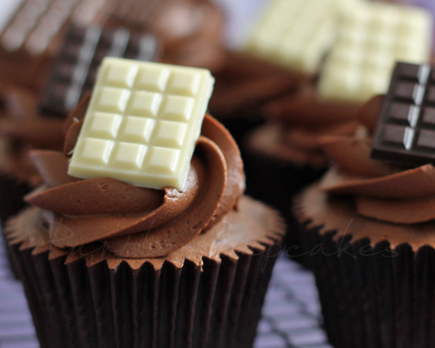 A close-up of chocolate cupcakes topped with swirls of rich chocolate frosting. Each cupcake is adorned with a piece of white chocolate or dark chocolate on top, adding a decorative touch. The cupcakes are presented in black liners, and the background features a blurred surface.