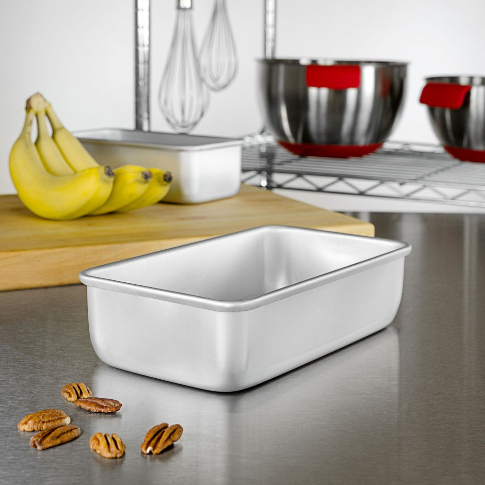 A stainless steel countertop with a shiny silver bread pan in the foreground, surrounded by scattered pecans. In the background, there is a wooden cutting board with three ripe bananas and a white bread pan. Behind this, two metallic mixing bowls with red handles and whisks hanging above them are visible.