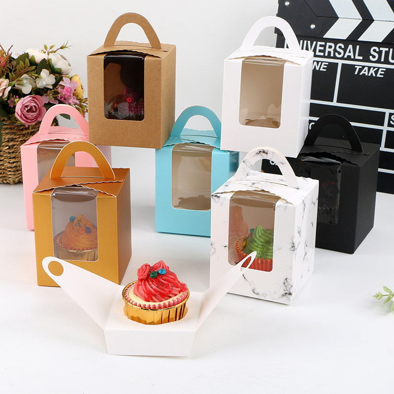 A collection of colorful cupcake boxes, designed for single portions, is displayed. The boxes are in various colors, including brown, white, black, light blue, pink, and marbled patterns, each with a clear front panel for viewing the cupcakes inside. In the foreground, there is a white cupcake holder displaying a cupcake with vibrant red frosting and decorative elements. The setting includes floral arrangements in the background along with a film clapperboard, adding a creative touch to the display.