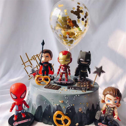 A decorated birthday cake featuring five colorful mini cartoon superhero figurines from the Avengers. The characters displayed include Iron Man, Spider-Man, a superhero with a spear, Batman, and a figure resembling Thor. The cake is gray with a textured surface and adorned with chocolate pieces, pretzels, and gold confetti. Above the cake, there is a clear balloon filled with gold confetti and a "Happy Birthday" sign made of gold lettering. The setting has a soft, light background.