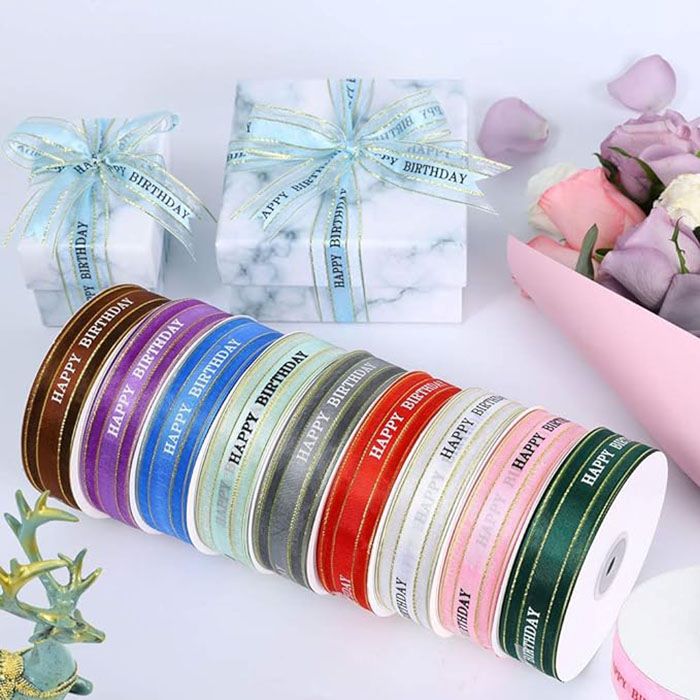 The image features a collection of colorful ribbons arranged neatly in a semi-circle. Each ribbon is about 2.5 cm wide and bears the text &quot;HAPPY BIRTHDAY&quot; in various colors, including shades of brown, purple, blue, pink, and green. In the background, there are two beautifully wrapped marble-patterned gift boxes adorned with matching ribbons. To the side, there is a bouquet of pastel-colored flowers partially visible. The overall presentation is festive and suitable for birthday celebrations.