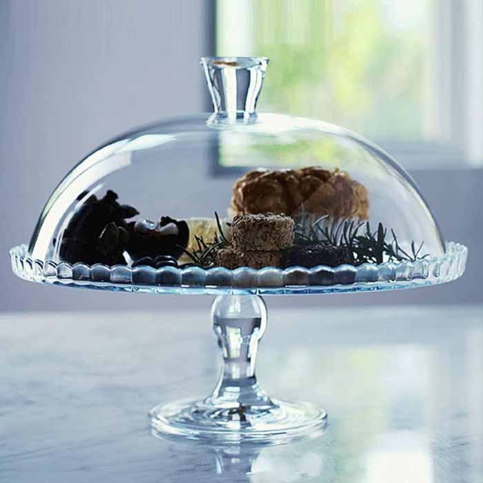 A clear glass cake display stand featuring a dome cover, showcasing a selection of desserts and treats. The stand has a decorative scalloped edge and a central pedestal base, with pastries and chocolate items arranged inside under the dome. The background is softly lit with natural light coming through a window.