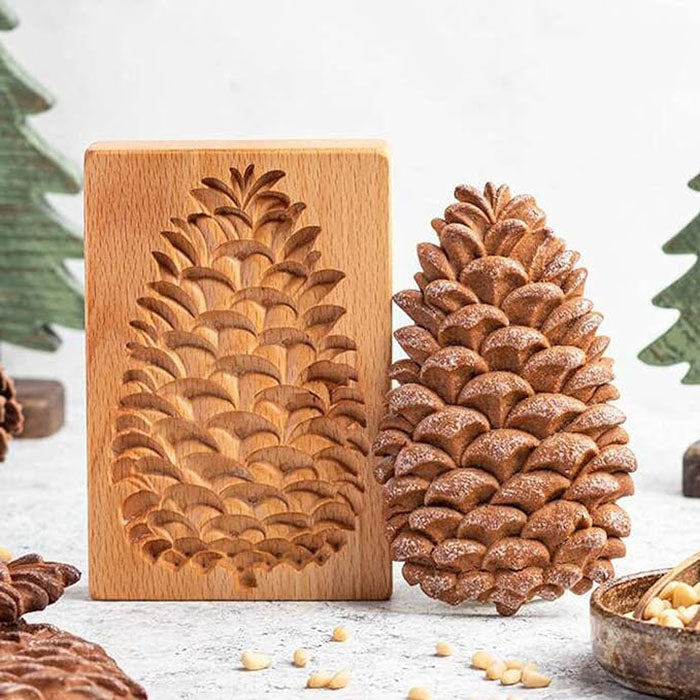 An image shows a pinecone-shaped silicone cookie mold alongside a freshly baked cookie that resembles a pinecone. The mold is made of light-colored wood with an intricate carved design of a pinecone on one side. The cookie, darker and detailed, has a textured surface mimicking the scales of a pinecone, dusted lightly with flour. In the background, there are decorative green trees and a small bowl with