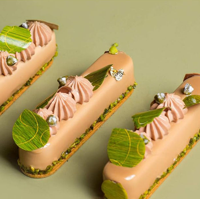 Three elegant eclair desserts arranged on a pale green surface. Each eclair is topped with pink creamy swirls and garnished with green leaves and small decorative elements. The base of each eclair is adorned with a layer of crushed nuts, adding texture and contrast to the smooth, glossy finish.