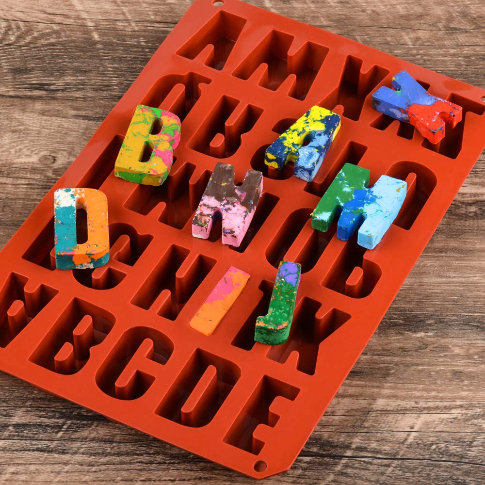 An orange silicone mould containing large uppercase letters is displayed on a wooden surface. Several colorful, painted letter-shaped objects are visible, some partially placed in the mould. The letters feature various bright colors and patterns, indicating they may be made of a different material such as plaster or crayon. The mould is designed for creating shaped letters, with clear outlines for each letter.