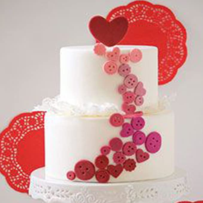 A two-tiered white cake is decorated with a cascading pattern of colorful buttons in shades of pink and red, creating a whimsical effect. At the top, a red heart-shaped decoration sits prominently. In the background, there are red lace-like doilies adding texture to the overall presentation. The cake is placed on an ornate, white cake stand.
