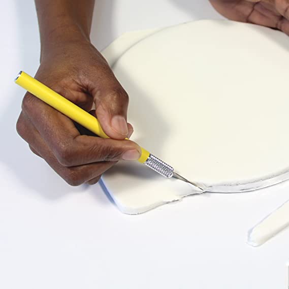 A close-up image shows a person using an art knife to cut into a white, round piece of material on a flat surface. The art knife has a yellow handle and a silver blade, while the person&