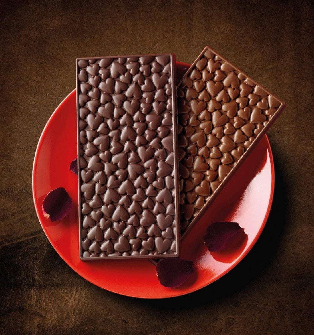 A red plate holds two chocolate bars with heart-shaped designs. The left bar is a darker chocolate, while the right bar is a lighter chocolate. Scattered rose petals are placed around the chocolate bars, and the background is a dark, textured surface, enhancing the romantic theme.