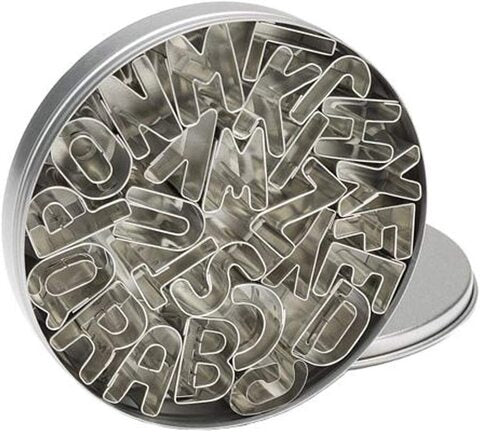 A round metal tin filled with various alphabet cookie cutters is shown. The cookie cutters are in the shape of letters, arranged closely together. The lid of the tin is slightly open, revealing the metal cutters, which are shiny and have various letter shapes, including multiple instances of some letters. The background appears to be a light color, complementing the metallic look of the cutters.