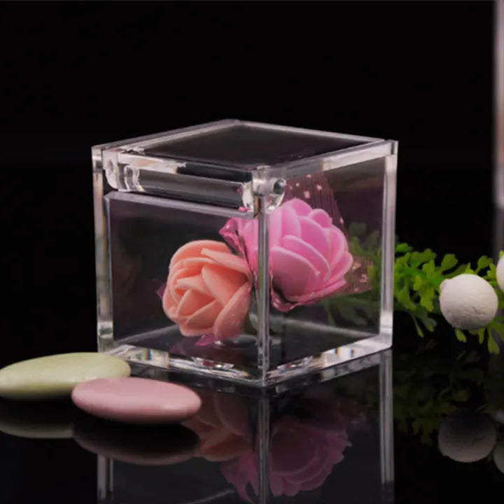 A clear acrylic cookie box is displayed in the image, partially filled with artificial flowers in shades of pink and peach. The box has a lid and features a shiny surface that reflects the surrounding environment. Next to the box, there are three decorative pastel-colored candies: one light green, one pink, and one white. The background is dark, enhancing the visibility of the box and its contents.