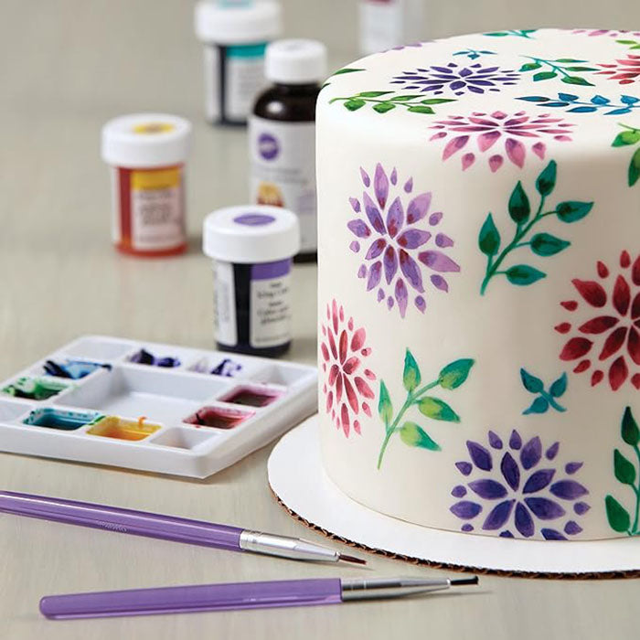 An image featuring a decorated white cake adorned with colorful floral designs in pink, purple, and green. In the foreground, there are two purple brushes, and a color palette with small wells containing various colored paints. In the background, several bottles of edible paint are visible. The setting appears to be a cake decorating workspace.