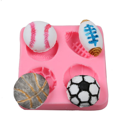 A pink silicone mold featuring four spherical cavities, each shaped like different sports balls. The top left has a white baseball with red stitching, the top right shows a white American football with blue detailing, the bottom left contains a grey basketball with orange lines, and the bottom right includes a black and white soccer ball.