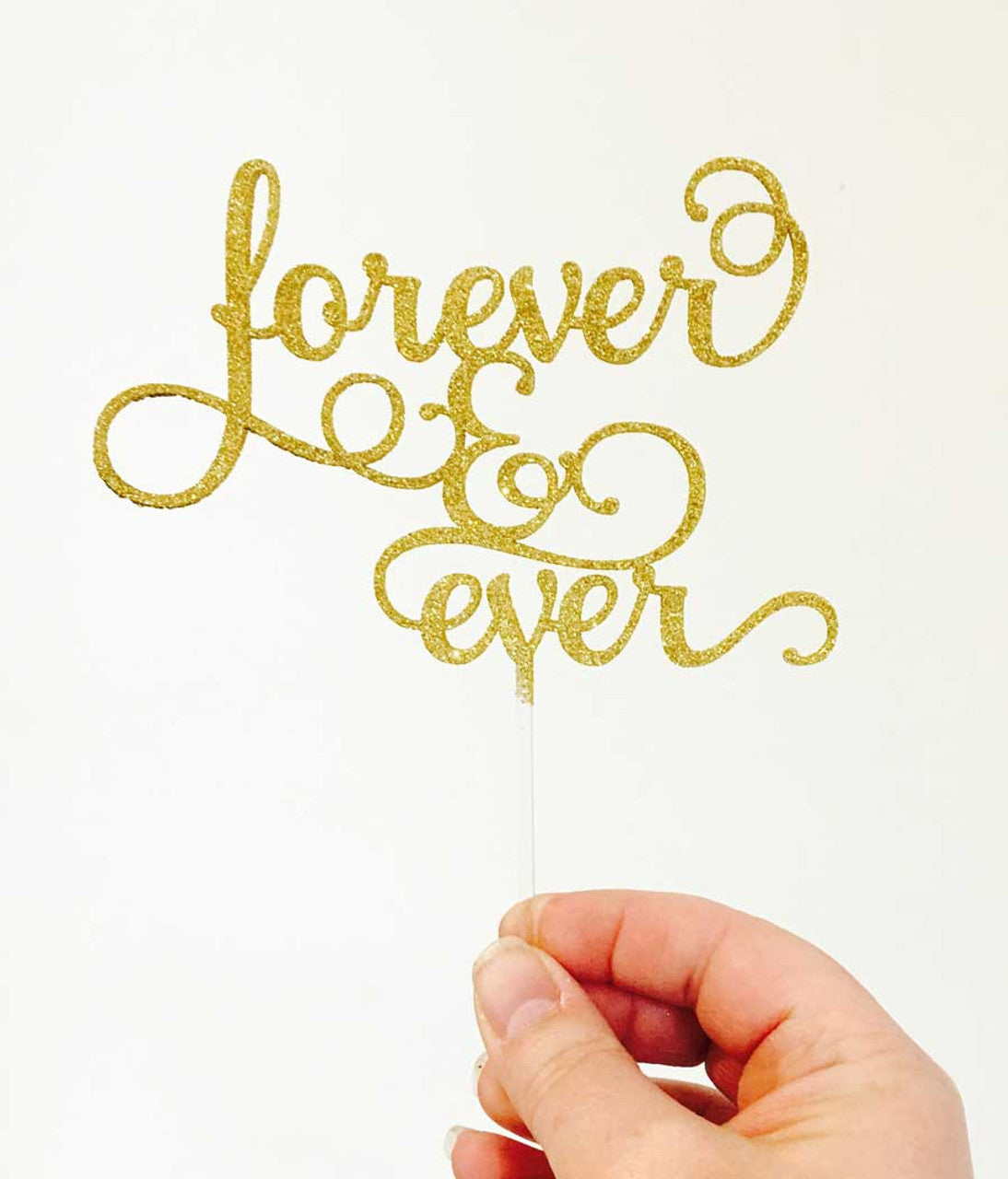 A hand holds a gold glitter acrylic cake topper that reads &quot;forever &amp; ever&quot; in a cursive font. The topper has a clear stick for insertion into a cake, and the background is plain white.