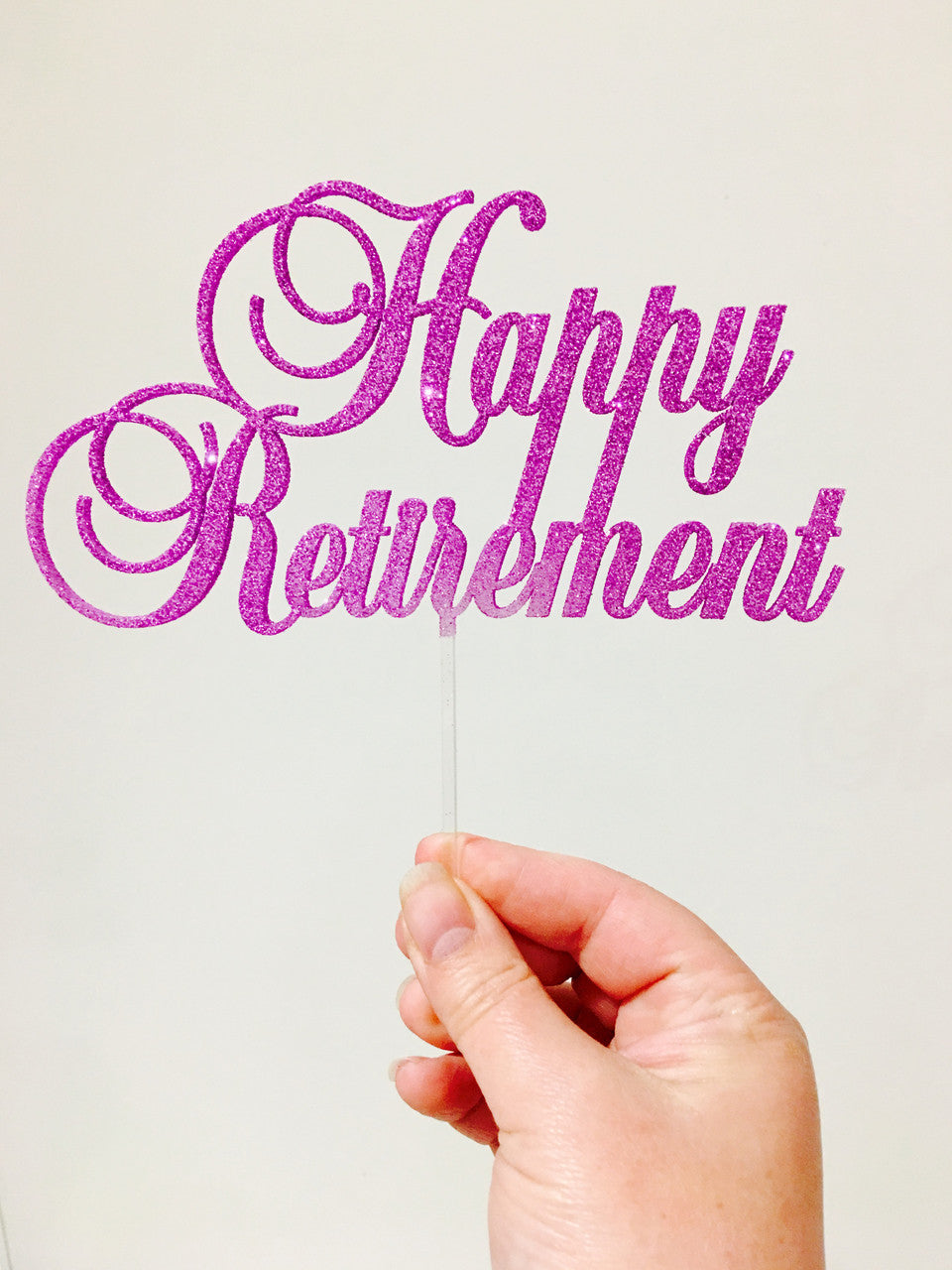 A hand holding a glittery pink acrylic cake topper that reads &quot;Happy Retirement.&quot; The topper features decorative cursive lettering and is attached to a clear stem. The background is a plain, light color.