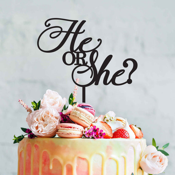 An elegant acrylic cake topper saying "He or She?" in stylish, swirling lettering is prominently displayed on top of a colorful drip cake. The cake features a pale yellow-orange icing with decorative flowers, strawberries, and macarons in shades of pink and purple surrounding the base. The background is softly blurred, focusing on the cake and topper.