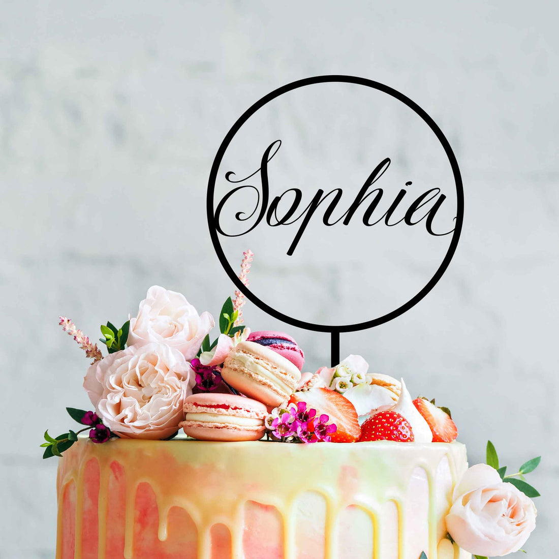 A beautifully decorated round birthday cake featuring a pink and yellow drip design. The cake is topped with a variety of colorful macarons, fresh strawberries, and white flowers, including pale pink roses. Above the cake, a black acrylic topper is displayed in a circular shape, with the name &quot;Sophia&quot; elegantly scripted inside. The background is softly blurred, enhancing the focus on the cake.