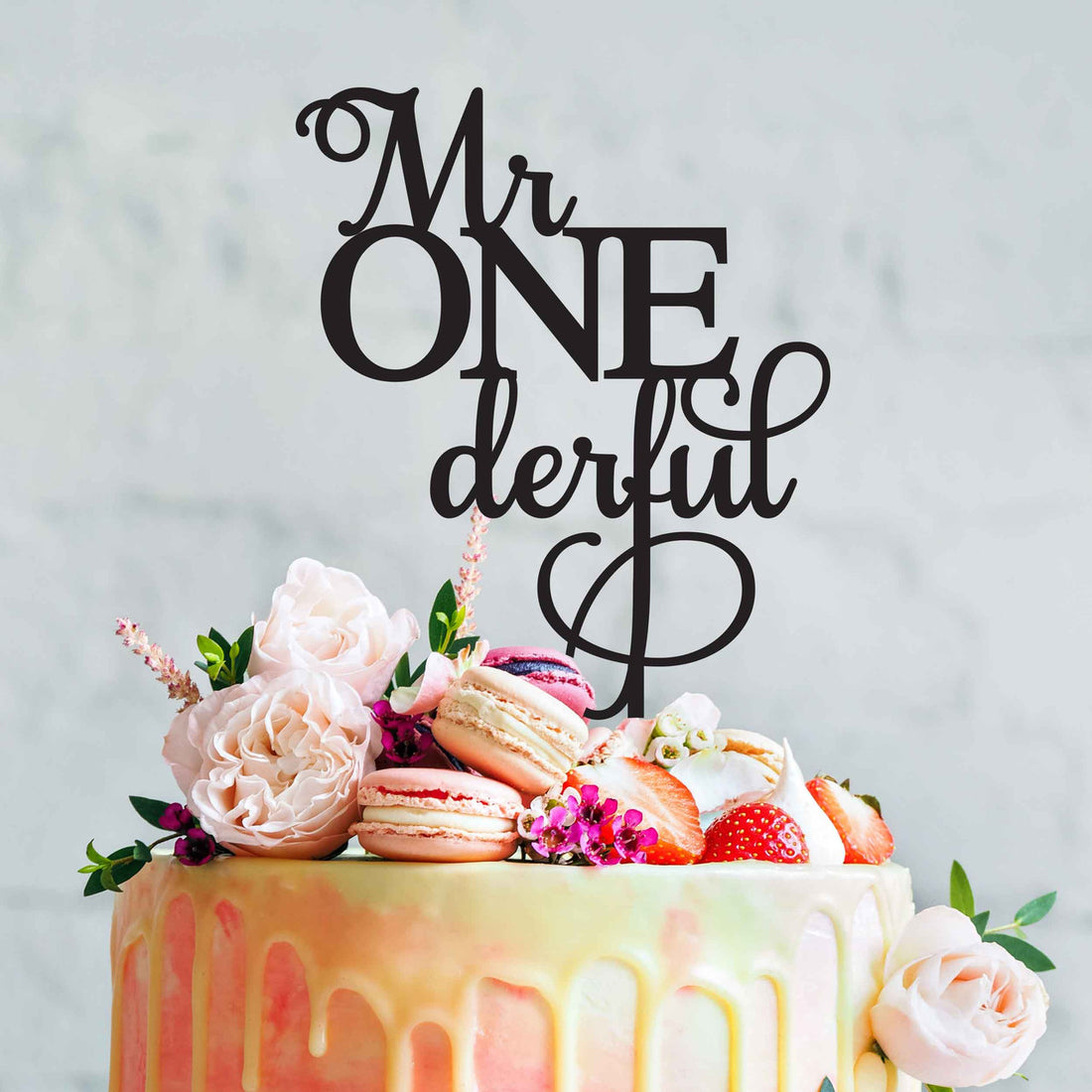 A beautifully decorated cake features an acrylic topper that reads "Mr ONederful" in elegant black lettering. The cake is adorned with pastel-colored icing drips and topped with an arrangement of fresh flowers, macarons, strawberries, and decorative elements. The background is a light, soft color.