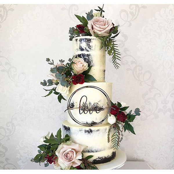 A three-tiered cake decorated with white and black icing. The cake features floral arrangements with pink roses, red roses, and greenery. A circular acrylic cake topper in the center displays the word &quot;love&quot; in an elegant script. The background has a subtle patterned design.