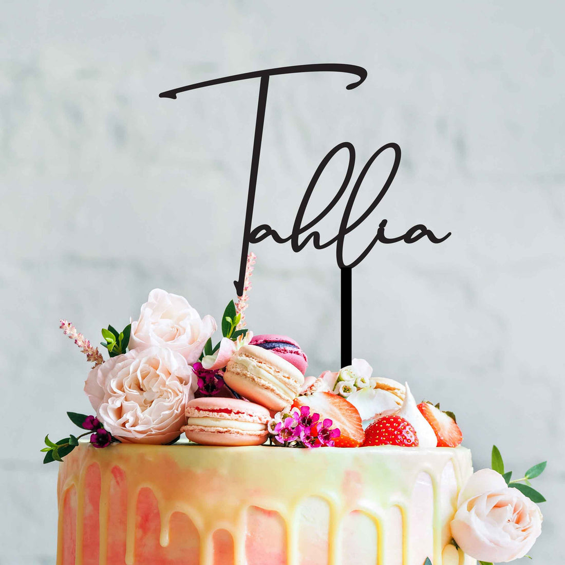 A beautiful birthday cake decorated with a custom acrylic topper that reads &quot;Tahlia&quot; in an elegant cursive font. The cake features a smooth, pastel-colored icing with a glossy drip effect at the edges. It&