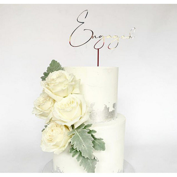 A two-tier white cake topped with a gold acrylic topper that reads &quot;Engaged.&quot; The cake features a decorative arrangement of white roses and green foliage on one side. The bottom tier has a subtle silver sheen at the base. The background is plain white.