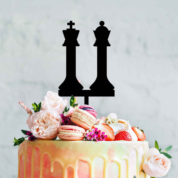 A colorful, decorated cake topped with a black acrylic cake topper shaped like king and queen chess pieces. The cake features pink and orange drip icing, along with various toppings including macarons, strawberries, and flowers. The background is a light, textured surface.