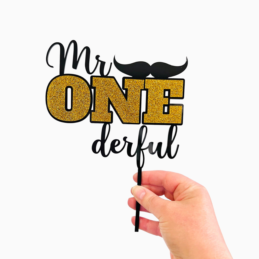 A hand is holding a cake topper that reads "Mr ONE derful." The word "Mr" is in a stylish font, while "ONE" is prominently displayed in bright gold glitter. Below "ONE," the word "derful" is in a cursive, black font. A small black mustache is positioned above the word "ONE." The overall design is festive and suitable for a birthday celebration, particularly for a first birthday. The backdrop is plain white.