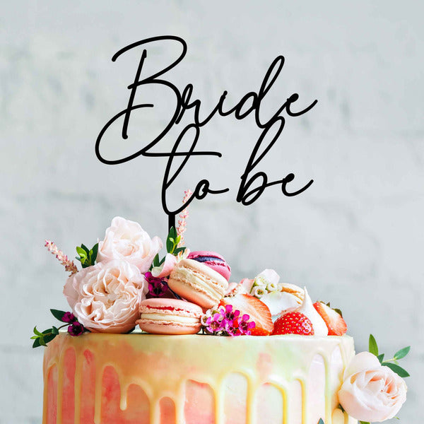 The image features a beautifully decorated cake adorned with various toppings. At the top, there is a stylish acrylic cake topper that reads &quot;Bride to be&quot; in elegant, cursive lettering. Surrounding the topper are pink and white flowers, colorful macarons, and fresh strawberries, adding a festive touch to the cake. The cake itself has a gradient frosting with a smooth, drippy design, set against a light, neutral background.