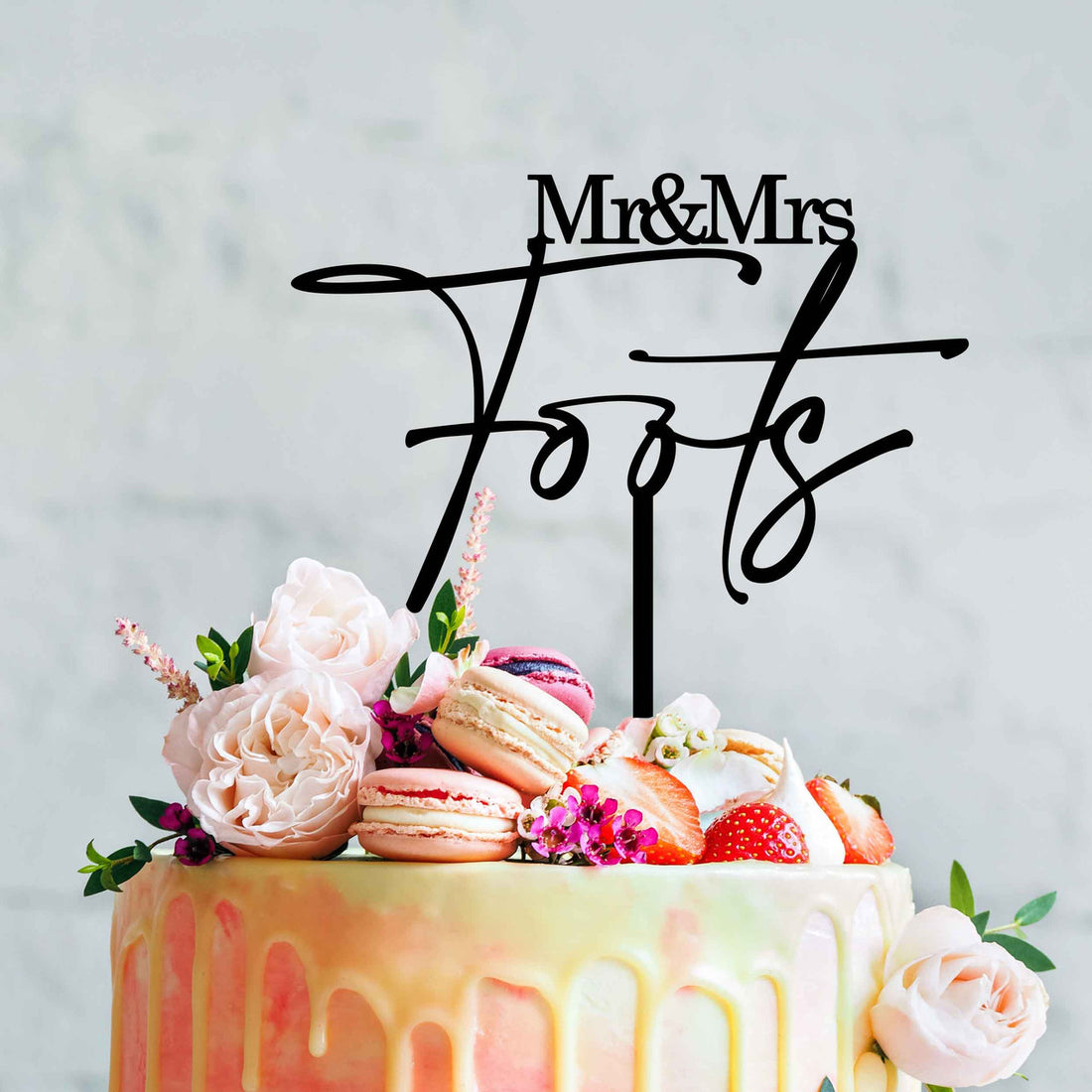 The image features a wedding cake topped with an acrylic cake topper that reads &quot;Mr &amp; Mrs&quot; followed by the surname &quot;Foots.&quot; The cake is decorated with a smooth, pastel-colored icing that has a glossy drip effect. Surrounding the cake topper are decorative elements including fresh flowers in shades of pink and white, macarons in various colors, and strawberries. The background is a soft, muted color that complements the cake&