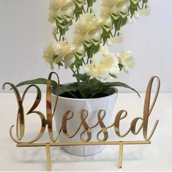 The image features a gold acrylic cake topper displaying the word &quot;Blessed&quot; in an elegant cursive font. The topper is set in front of a white pot containing a bouquet of soft, white artificial flowers with green leaves. The background is neutral, enhancing the visibility of both the cake topper and the floral arrangement.