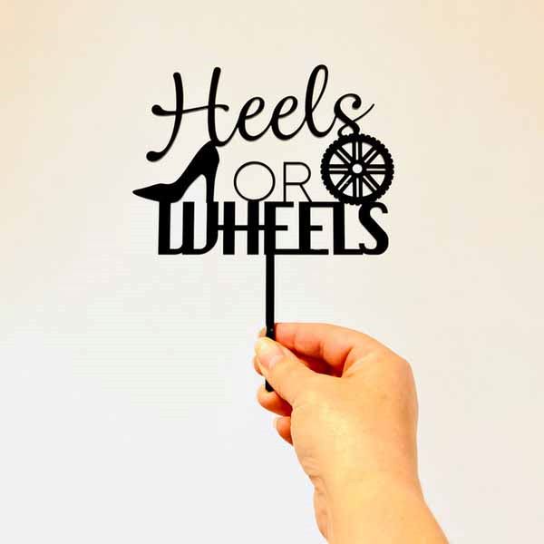 A hand is holding a black acrylic cake topper that features the words "Heels or WHEELS." The design includes a stylized high heel shoe next to the word "Heels" and a wheel icon next to the word "WHEELS." The background is plain and neutral in color, allowing the topper to stand out prominently.