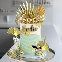 A decorated cake featuring a smooth, light blue base and white frosting. The cake is topped with an arrangement of acrylic gold ginkgo leaves and other decorative elements, including golden beads and greenery. The name &quot;Sofia&quot; is displayed in an elegant script on the cake&