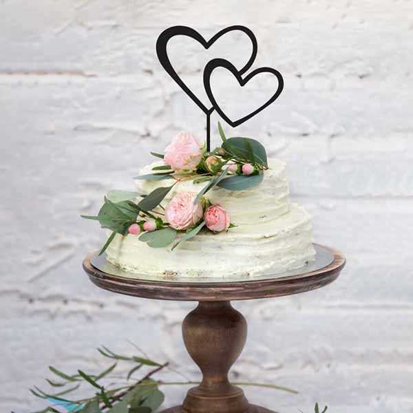 A two-tiered, frosted cake sits on a wooden cake stand. Atop the cake, there are two overlapping black heart-shaped cake toppers. The cake is decorated with fresh pink flowers and greenery, adding a romantic touch. The background features a light-colored, textured wall.