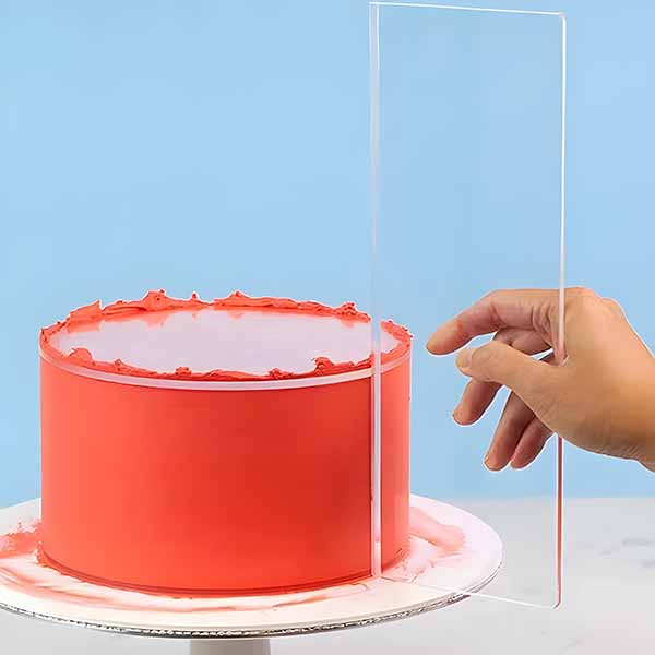 A hand holds a clear acrylic cake scraper next to a round cake that is frosted in red icing, with some excess icing visible on the base. The background is a light blue color, and the cake sits on a white, circular cake board. The scraper appears to be used for smoothing the cake&