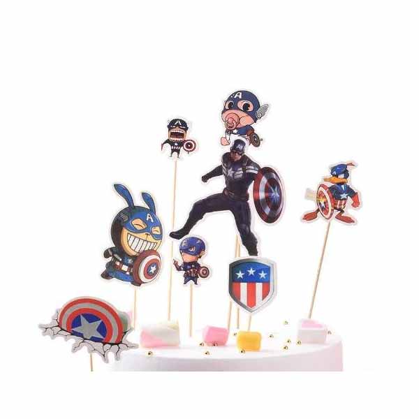 A colorful cake topper featuring various Captain America-themed decorations. Central is a figure of a superhero with a shield, surrounded by cartoonish characters in superhero costumes, including a bunny and other playful interpretations of Captain America. There are also small decorative shields and cake elements like marshmallows and edible stars on a white cake base.