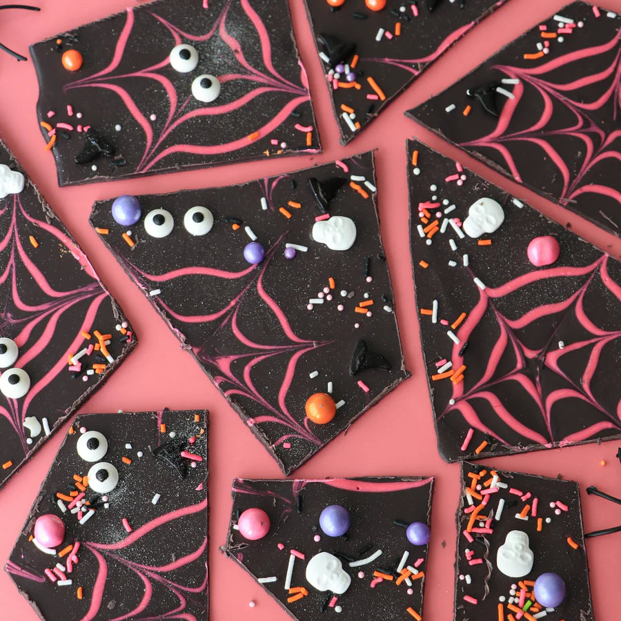 The image features several pieces of dark chocolate bark, each with a decorative design. The bark is adorned with pink spider web patterns and various colorful sprinkles, including orange, purple, and black. There are also small decorations shaped like eyes and skulls scattered across the surface. The background is a soft pink, enhancing the vibrant colors of the toppings.