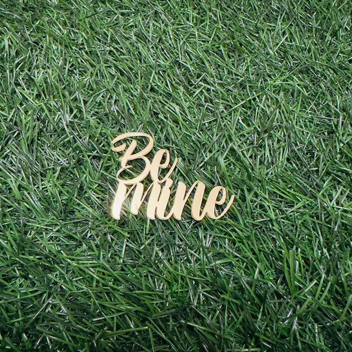 A gold, cursive cake topper that reads "Be Mine," positioned on a green artificial grass background.