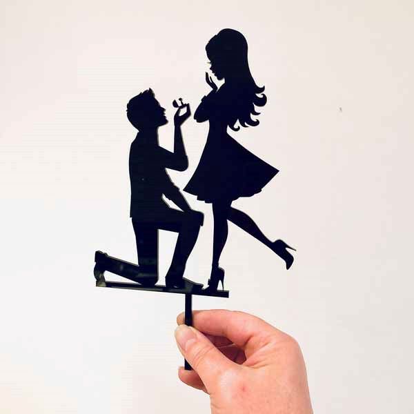 The image features a hand holding a black acrylic wedding cake topper. The topper depicts a silhouette of a man kneeling on one knee, proposing marriage with an engagement ring. In front of him stands a woman with a playful pose, wearing a dress and holding her hands to her face, suggesting surprise or delight. The overall design is elegant and emphasizes the romantic theme of a marriage proposal.