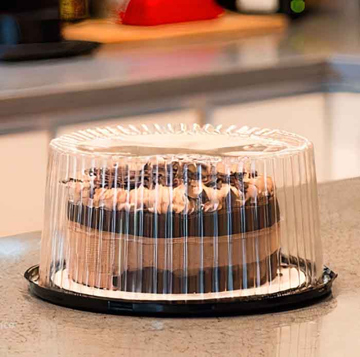 A black base round cake container with a clear, ribbed plastic lid is displayed on a kitchen countertop. Inside the container, a cake is visible, featuring dark frosting and decorative elements on top. The setting appears to be a kitchen, with soft lighting in the background and various kitchen items partially visible.