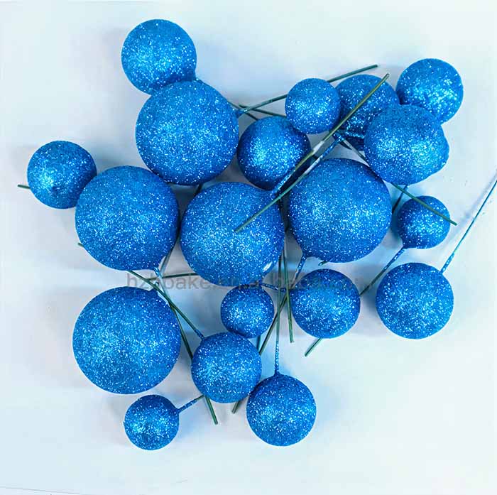 A collection of blue glittery balls in various sizes, some attached to green wire stems. The balls are arranged closely together on a white surface, creating a vibrant and decorative appearance suitable for cake decoration.