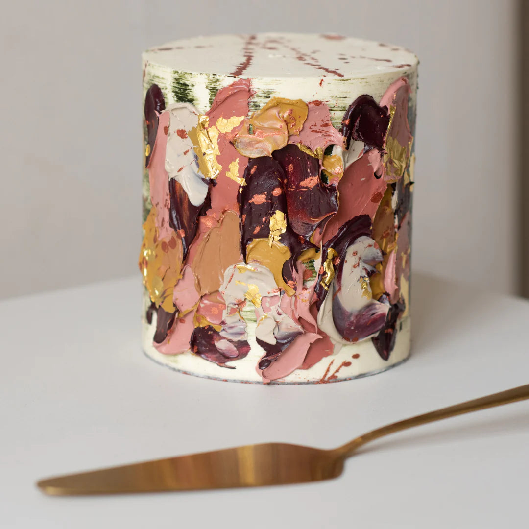 A decorative cake with various shades of pink, brown, and cream frosting, adorned with gold leaf accents. The cake has a textured appearance with swirls and layered colors. A gold-colored cake knife is placed beside it on a light-colored surface.