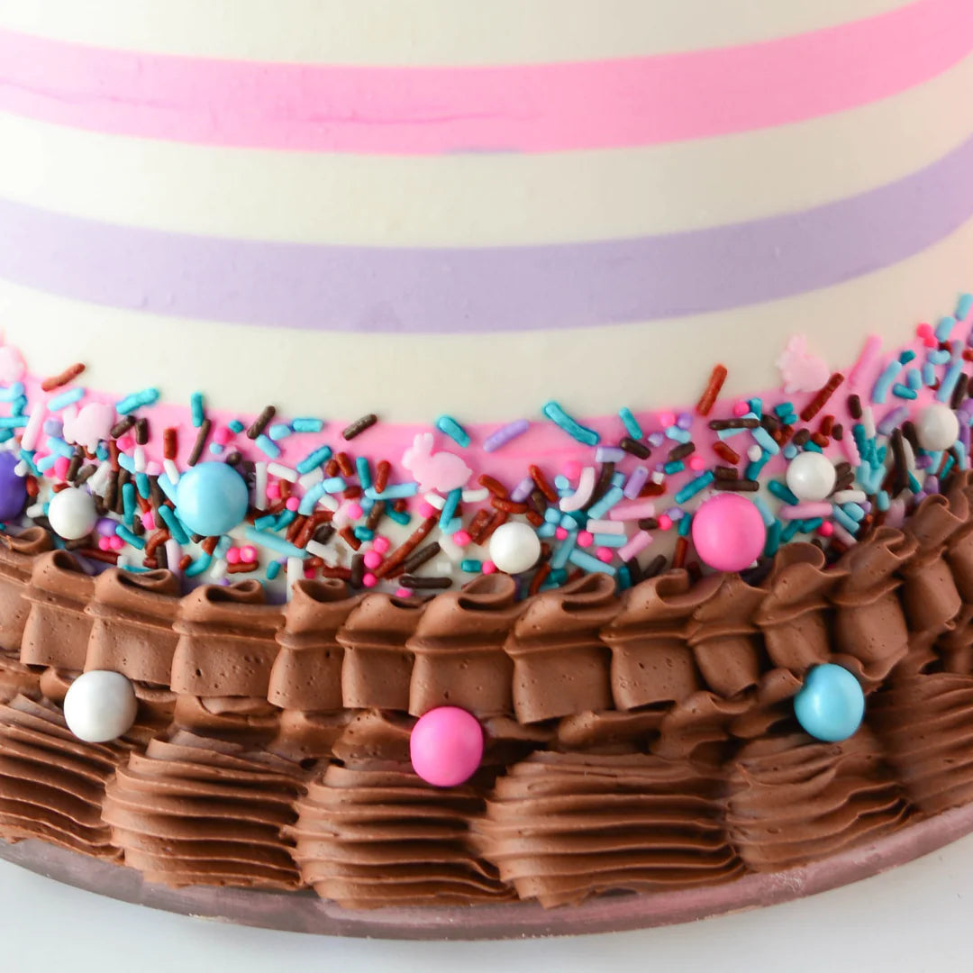 The image shows a decorated cake with a smooth, light-colored frosting featuring horizontal pink and lavender stripes. The base of the cake is adorned with a layer of chocolate frosting, intricately piped in waves. Surrounding the chocolate frosting are colorful sprinkles in various shapes and sizes, including pink, blue, and white candies, as well as chocolate sprinkles, creating a vibrant and festive appearance.